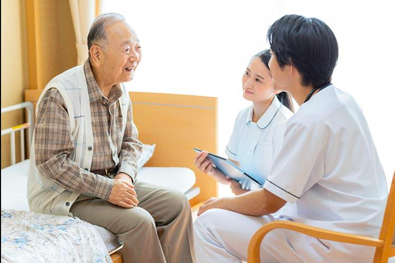 居室のベッドに座る利用者と、タブレットを手に話を聞く看護師・スタッフ