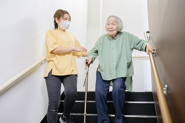 階段で杖をつき、スタッフに付き添われて歩行訓練をする利用者