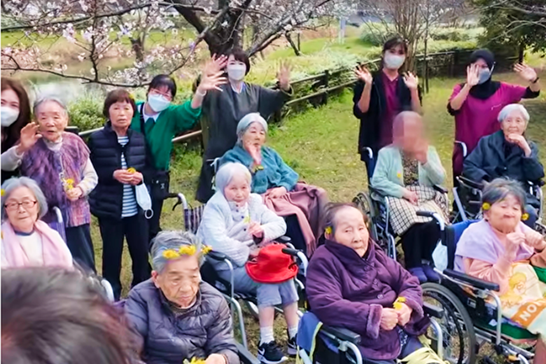 桜の下で車いすの利用者とスタッフが集まり、手を振るお花見の様子